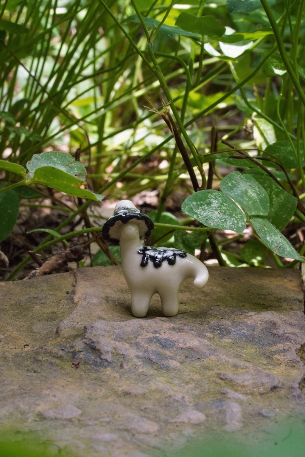 Small figurine of a white animal wearing a black hat on a stone surface with green foliage in the background